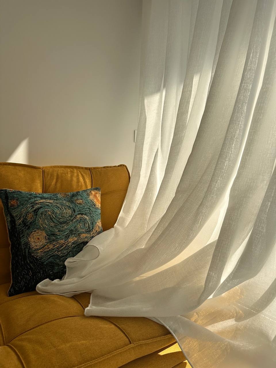 Detalle de visillo blanco de lino cayendo junto a un sofá amarillo con cojín