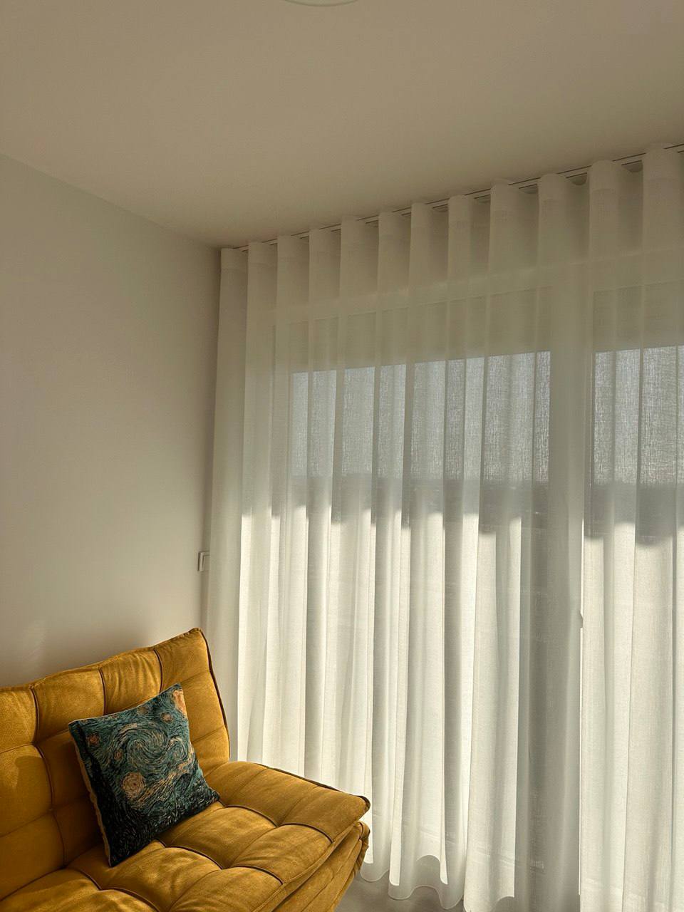 Visillos blancos de caída ondulada filtrando la luz sobre un sofá amarillo