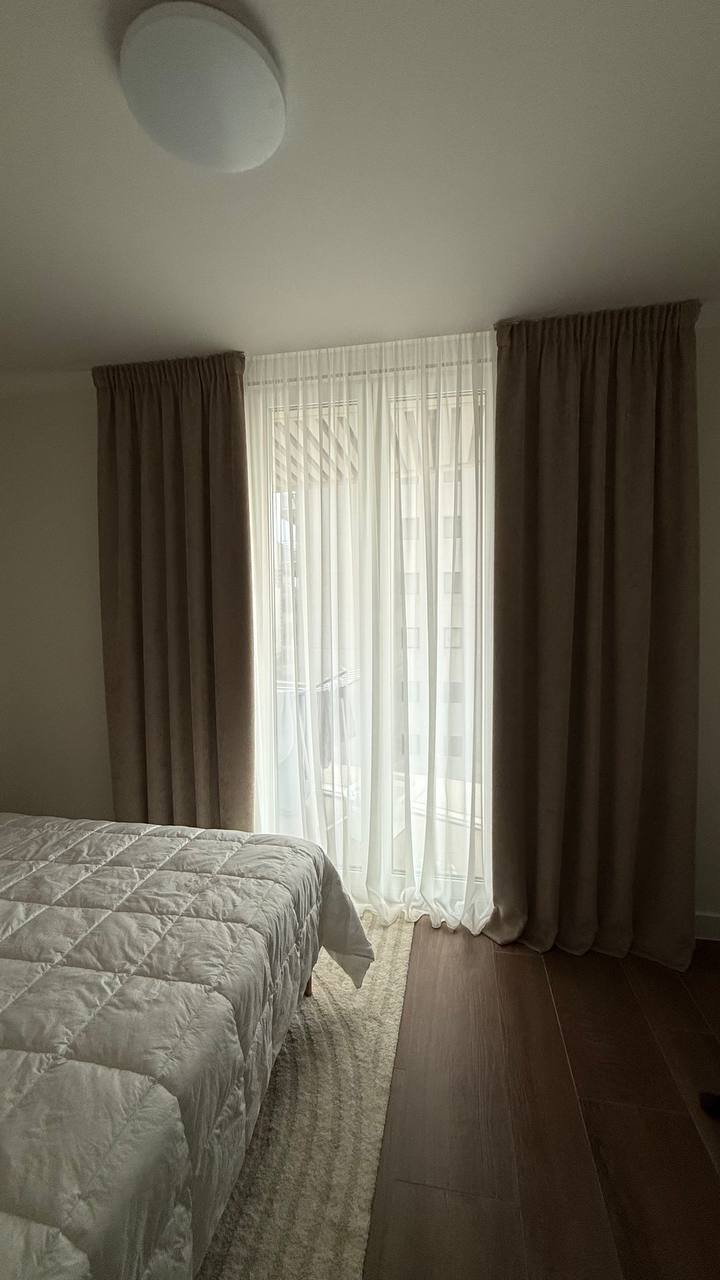 Dormitorio con cortinas beige y visillo blanco en el ventanal del balcón